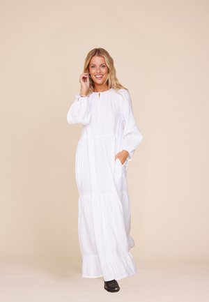 Kvinne i en lang hvit kjole med lag og lange ermer, smilende, med den ene hånden som berører håret og den andre i kjolens lomme.