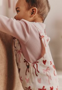 Bela kombinezona z rdečimi in roza vzorci src in pentelj, oblečena čez svetlo rožnato rebrasto majico z dolgimi rokavi, na teksturirani rjavi podlagi.