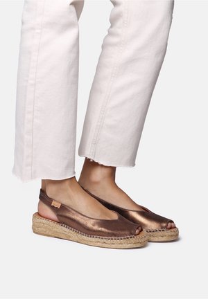 Fødder iført bronze-metalliske åbne slingback-espadriller kombineret med off-white cropped bukser med frynsete kanter.
