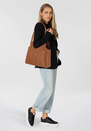 Femme portant un blazer noir, un jean roulé bleu clair, des baskets noires, portant un grand sac fourre-tout en cuir brun avec un design perforé sur l'épaule.