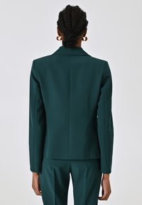 Blazer verde scuro su misura con una silhouette strutturata, tessuto morbido, rever affilati e un'unica fessura posteriore, mostrato da dietro.