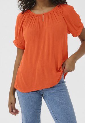 Femme portant un chemisier à manches courtes orange vif avec un col froncé et un jean bleu, la main reposant dans la poche, fond neutre.