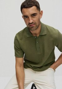 Oliwkowa koszulka polo o fakturowanym wykończeniu, z krótkimi rękawami, trzema guzikami z przodu i klasycznym kołnierzykiem, w połączeniu z jasnymi spodniami.