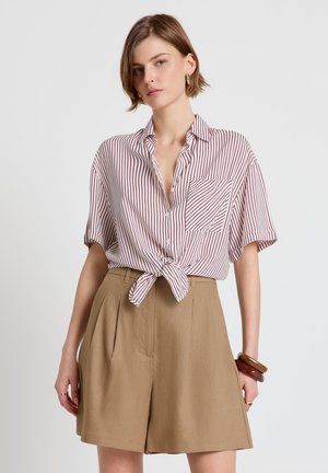 Femme portant un short taille haute beige et une chemise blanche à manches courtes avec des rayures rouges nouée à la taille, accessoirisée avec des bracelets et des bagues.