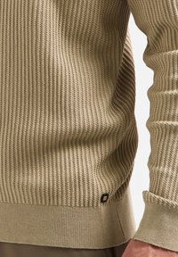 Beige geribbelde trui met een gestructureerde oppervlakte, voorzien van een ronde hals en omgeslagen mouwen. Een klein, zwart en gouden logo is zichtbaar aan de zoom.