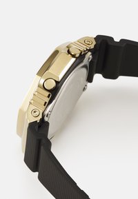 Montre dorée avec un bracelet en caoutchouc noir, dotée d'un boîtier rectangulaire, d'accents texturés et de trois boutons latéraux pour la fonctionnalité.
