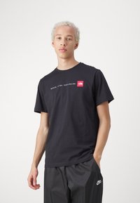 Zwarte katoenen t-shirt met witte tekst "NEVER STOP EXPLORING" en een rood logo patch. Ronde hals en korte mouwen.