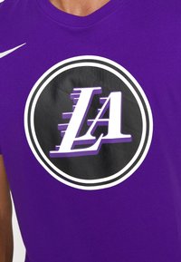 T-shirt en coton violet avec un emblème noir circulaire comportant un design "LA" blanc et violet, accentué par une bordure blanche et un logo Nike.