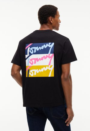 Mann trägt schwarzes T-Shirt mit buntem rechteckigem grafischem Motiv und stilisiertem, wiederholtem Text auf der Rückseite, steht vor einem schlichten weißen Hintergrund.
