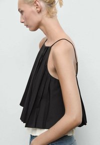 Donna con i capelli biondi corti che indossa un top senza maniche nero plissettato con spalline sottili e jeans blu, rivolta verso sinistra su uno sfondo neutro.