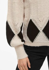 Pletený svetr v světle růžové barvě s černými diamantovými nášivkami na rukávech. Má žebrované manžety a jemné vzory po celém svršku.