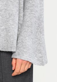 Szary sweter z miękkiego, teksturowanego materiału. Charakteryzuje się szerokimi rękawami i luźnym krojem, zestawiony z wzorzystym czarnym materiałem u dołu.