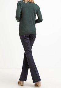 Mörkgrön tröja med rund halsringning, matchad med marinblå utsvängda byxor. Tan högklackade skor fulländar outfiten. Texturen ser stickad ut.