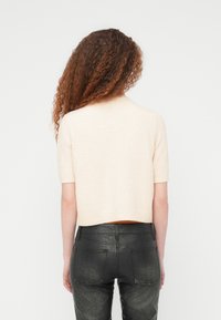 Γυναίκα με σγουρά μαλλιά που φοράει μπεζ πουλόβερ με κοντά μανίκια και μαύρο ψηλόμεσο παντελόνι, γυρισμένη πλάτη μπ ροστά σε απλό φόντο.