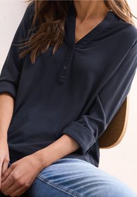 Chemise bleu marine à manches longues avec un col en V et trois boutons. Tissu texturé, manches retroussées et coupe décontractée. Portée avec un jean bleu clair.