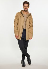 Lichtbruine jas met een capuchon, ritssluiting en zijzakken; gedragen over een beige shirt en gecombineerd met zwarte broeken en laarzen.