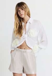 Weißes Hemd mit Rundkragen und strukturierten, floralen Applikationen, kombiniert mit hellen, metallischen Shorts. Lässiger, lockerer Schnitt mit leichtem Glanz.