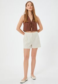 Top court marron avec un col en V et un détail à nouer, associé à un short beige clair. Le modèle porte des chaussures plates de couleur crème. Design simple.