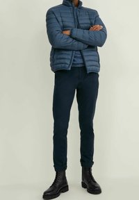 Veste matelassée bleu foncé avec fermeture éclair à l'avant, col côtelé, portée avec un pantalon bleu marine et des bottes noires à lacets. Texture lisse et matelassée.