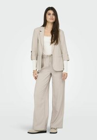 Blazer in lino beige e pantaloni a gamba larga coordinati, sovrapposti a una maglietta bianca a maniche lunghe. Tessuto morbido, vestibilità rilassata, con vita regolabile.