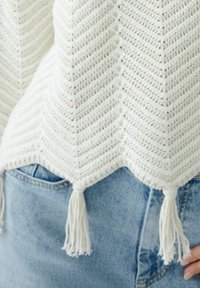 Weißer gehäkelter Poncho mit Zickzackmuster, gewelltem Saum und Quastenakzenten am unteren Rand, getragen über blauer Jeans.