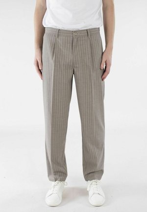 Person iført beige bukser med tynne striper og legg, og hvite joggesko, stående mot en ensfarget hvit bakgrunn.