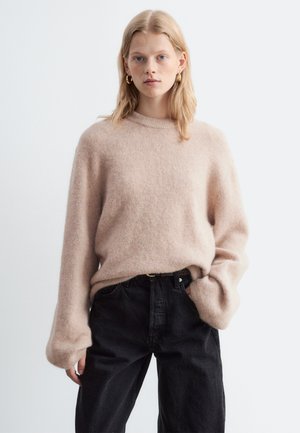 Jeune femme aux cheveux blonds portant un pull beige duveteux et un jean noir taille haute, posant avec une main dans la poche sur un fond uni.