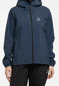 Veste imperméable bleu marine avec fermeture éclair à l'avant, capuche ajustable et poignets élastiques. Dispose d'un logo sur la poitrine et de deux poches latérales.