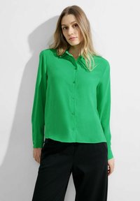 Camicia verde button-up realizzata in un tessuto morbido, con colletto e maniche lunghe, abbinata a pantaloni neri.