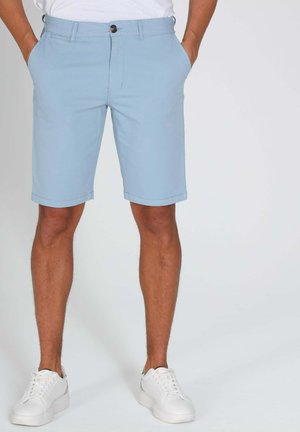 Homme portant un short bleu clair jusqu'aux genoux avec des poches avant, une boutonnière à boutons et une braguette à glissière, associé à des baskets blanches.