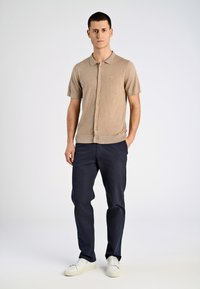 Beige kortärmad polo med knappar och krage, kombinerad med mörk marina byxor och vita sneakers. Mjuk textur, minimal detaljering.