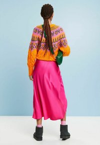 Pull orange en tricot avec motifs roses et violets, jupe en satin rose et bottines noires, avec sac bandoulière vert et cheveux tressés.