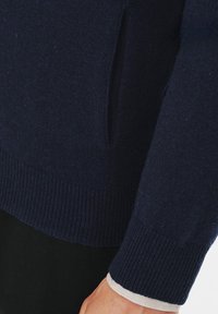 Granatowy sweter dzianinowy z ribbą na dolnym końcu i mankietach, z detalem bocznej kieszeni i miękką teksturą. Idealny do noszenia na warstwy.