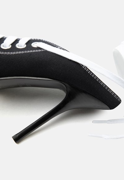 Chaussure noire à talon haut avec dessus en toile, lacets blancs, œillets métalliques et coutures blanches visibles, présentée sur un fond uni.