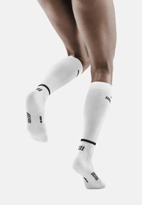 Calze compressive bianche con una texture a costine, fasce nere di accento sulla parte superiore e dettagli con logo sui lati e sulle suole.