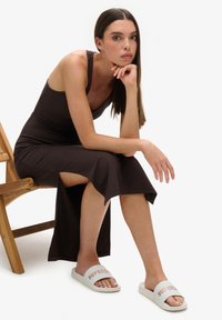 Brown sleeveless maxi dress with side slit, paired with white slide sandals featuring orange text. Textured wooden chair in the background.