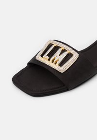 Sandale à glisser en suede noir avec une grande boucle rectangulaire dorée ornée de strass transparents. Texture lisse et design à bout carré.