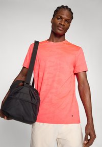 Camiseta de camuflaje rosa con cuello redondo y mangas cortas, combinada con un bolso de viaje negro texturizado que cuenta con una correa para el hombro.