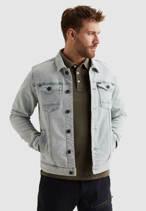 Mann mit kurzen braunen Haaren, trägt eine hellblaue Jeansjacke über einem olivgrünen Poloshirt, Hände in den Jackentaschen, blickt nach rechts.