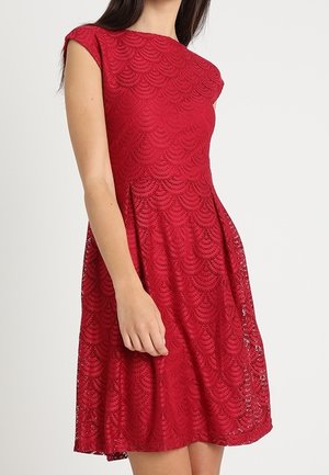 Femme portant une robe rouge en dentelle longueur genou avec des manches courtes et une taille cintrée sur fond blanc uni.