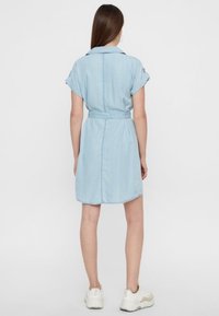 Kvinna i en ljusblå denimklänning som når knäet, med korta upprullade ärmar och bälte, kombinerad med vita sneakers, står vänd bort.