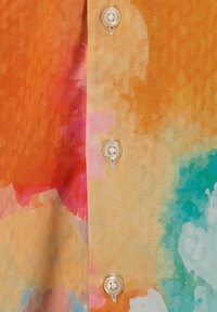 Nahaufnahme eines bunten Stoffes mit Mustern in Orange, Rot, Pink und Türkis sowie einer Reihe von weißen Knöpfen, die auf einer Knopfleiste genäht sind.