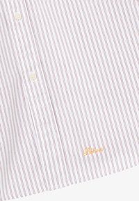 Weißes Hemd mit dünnen roten vertikalen Streifen, zwei weißen Knöpfen und gelb gesticktem Text "Błonie" in der Nähe des unterem Saums.