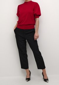 Haut rouge à manches courtes bouffantes, associé à un pantalon noir fuselé et des escarpins noirs à bout pointu avec des accents métalliques.