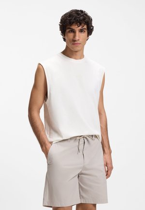 Jeune homme aux cheveux bouclés portant un débardeur blanc et un short beige à cordon, se tenant les mains dans les poches devant un fond blanc.