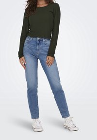 Frau steht mit den Händen an den Seiten, trägt ein olivgrünes Langarmshirt, hellblaue Jeans und weiße hohe Sneaker.