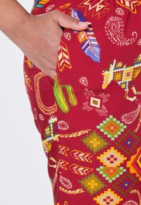 Un tessuto rosso con motivi presenta colorati disegni geometrici e di piume. Una mano si appoggia nella tasca laterale, evidenziando la texture vibrante del tessuto.