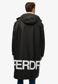 Abrigo negro impermeable con capucha, presenta el texto blanco "PERDR" en la parte posterior, tejido texturizado y un cuello forrado de forro polar.