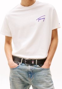 Mężczyzna w białej koszulce z krótkim rękawem z logo „Tommy”, jasnoniebieskie dżinsy i czarny pasek z prostokątną srebrną klamrą, ręce w kieszeniach.