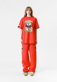 Camiseta roja de gran tamaño con un gráfico de oso de peluche, combinada con pantalones sueltos rojos a juego con acentos de oso de peluche en los bolsillos. Diseño casual.